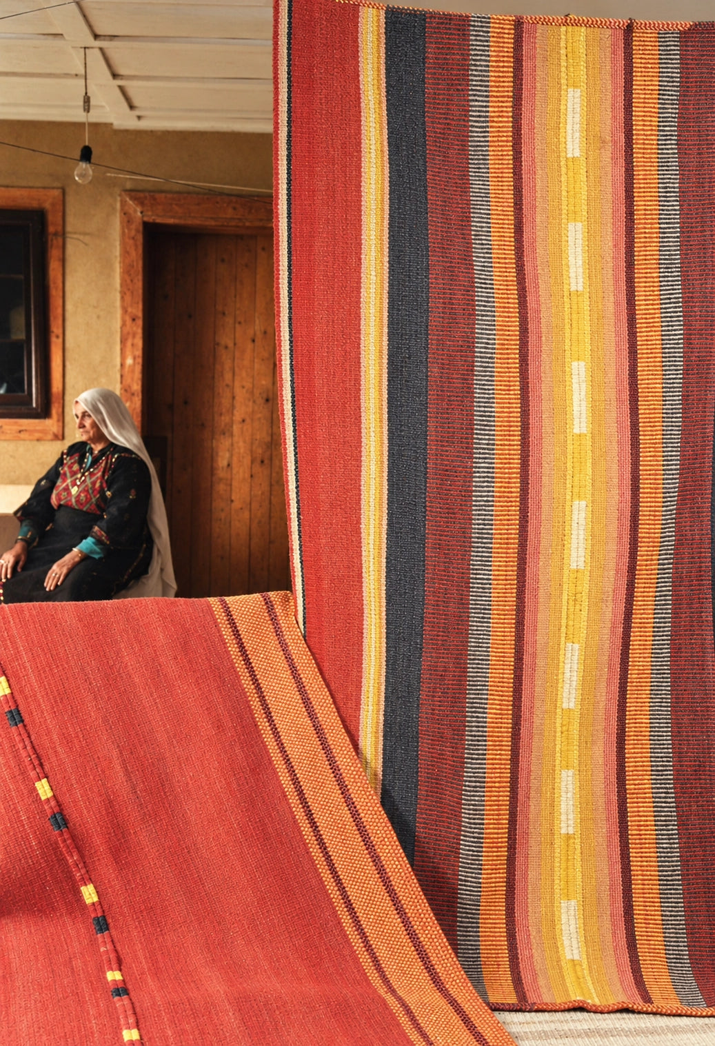 A woman in traditional clothing sits in a rustic room while large woven rugs are displayed, including a prominent red rug in the foreground and a tall striped rug with red, yellow, orange, black, and white bands.
