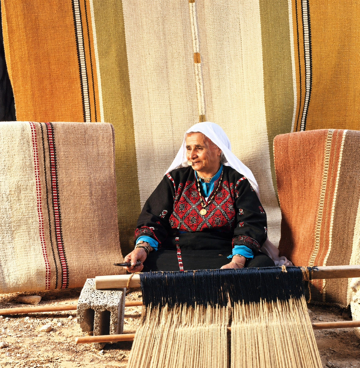 An Palestinian elderly woman wearing traditional clothing sits behind a handloom, actively weaving a striped textile. The background features other completed woven textiles in warm earthy tones of beige, brown, and orange.